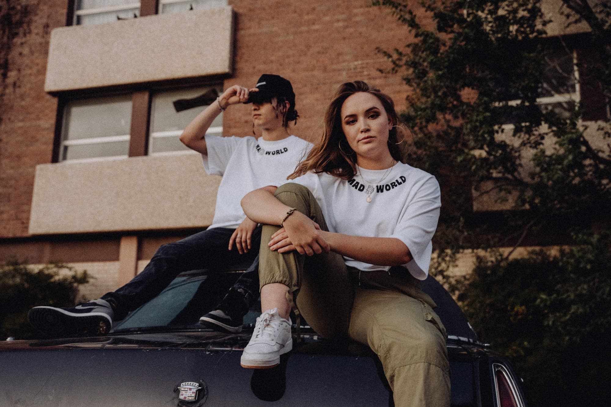 Two people sitting on the roof of a car outside a brick building, both in oversized white T-shirts