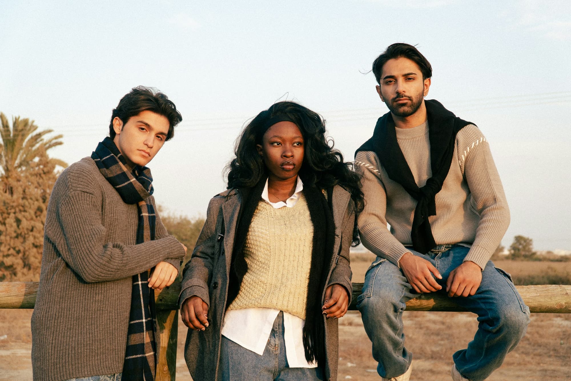 Three people leaning on a wooden fence at golden hour, in knitwear, a scarf and a long coat